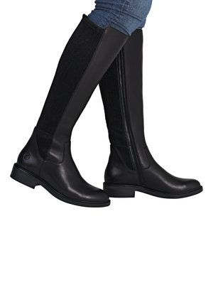 Black knee-high leather boots with ribbed elastic panels worn with blue denim jeans, shown from side angles on white background.