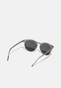 Sonnenbrille mit runden, dunklen Gläsern und transparenten grauen Rahmen. Schlanke Bügel mit leichter Krümmung, die ein minimalistisches Design zeigen.