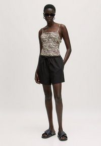 Zebra print camisole met dunne bandjes, gecombineerd met zwarte shorts met trekkoord. Model draagt zwarte zonnebril en slippers.