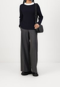 Frau trägt einen marineblauen Pullover, graue gestreifte Weitbeine-Hosen, schwarze klobige Schuhe und eine schwarze Umhängetasche mit einem markierten Gurt.