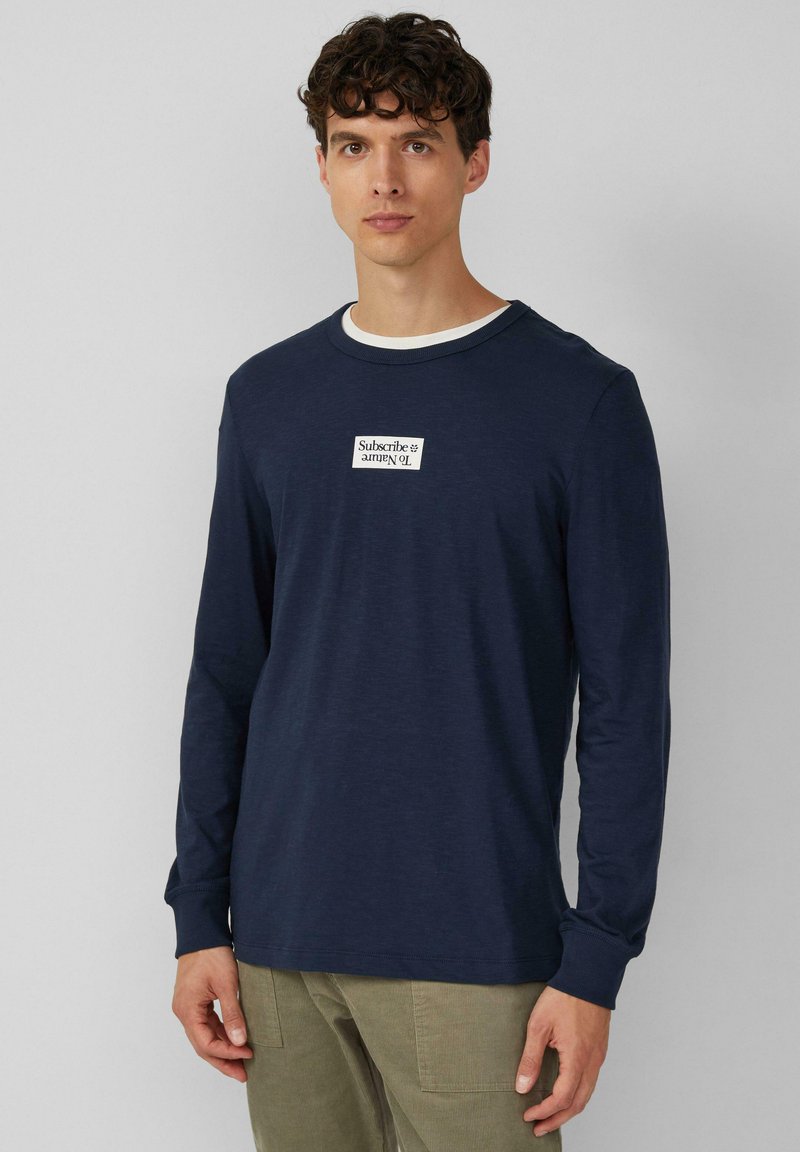 Maglietta a maniche lunghe blu navy con collo tondo bianco, caratterizzata da un'etichetta rettangolare bianca con testo nero sul petto. Texture morbida.