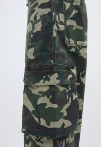 Kamo krāsas cargo bikses zaļos un melnos toņos, ar sānu kabatu un izturīgu audumu ar redzamām šuvju detaļām.