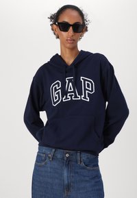 Marinblå hoodie med vit "GAP"-text, dragsko på huven och ficka framtill, paired med högmidjade blå jeans.