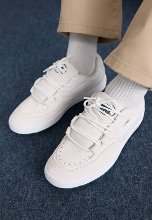 White suede sneakers with mesh accents, textured laces, and perforated details. The sole is light with a hint of turquoise. Worn with beige pants.