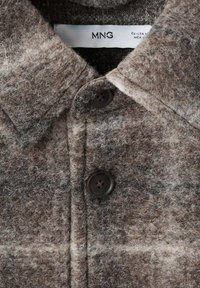 Cămașă din lână texturată de culoare maro, cu guler, având două nasturi negri și un model discret de carouri integrat în țesătură.