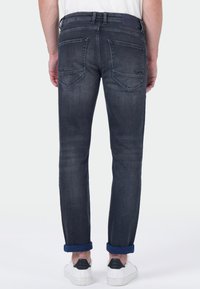 Man in donkere grijze opgerolde jeans en witte sneakers, staand met zijn rug naar de camera tegen een lichte achtergrond.