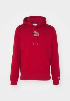 Sweat à capuche rouge avec logo Lacoste sur le devant et emblème crocodile vert sur le poignet de la manche gauche.