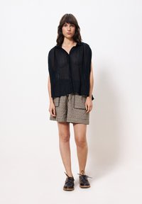 Schwarzes, durchsichtiges Kurzarmhemd mit Kragen, kombiniert mit einem gemusterten karierten Rock. Das Model trägt schwarze Schnürschuhe. Minimalistischer Hintergrund.