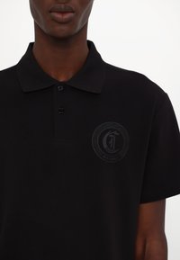 Svart poloskjorte laget av teksturert stoff, med en svart-på-svart logo med "JUST CAVALLI" på brystet og tre knappåpning.