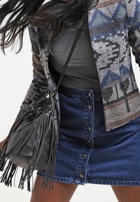 Femme portant une veste à motifs, un haut gris, une jupe en denim boutonnée, et portant un sac à main en cuir noir avec des franges sur son épaule.