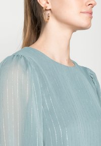 Top de tecido azul-claro com riscas verticais prateadas e mangas bufantes. Brinco de argola dourada visível. Textura suave e transparente.