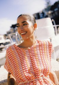 Femme souriante aux cheveux foncés, vêtue d'une robe à motifs rose, orange et rouge, assise en plein air par une journée ensoleillée près de bateaux et de l'eau.