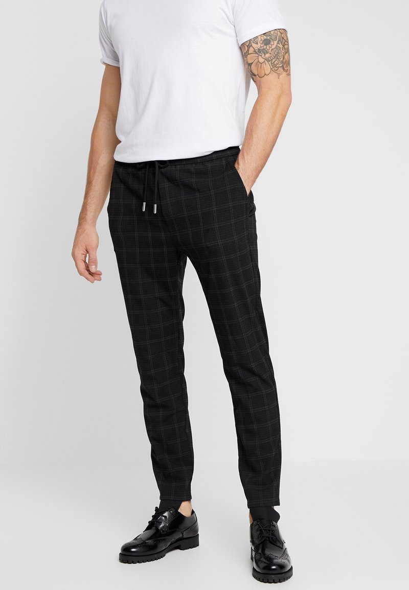 Svarta rutiga joggerbyxor med elastisk midja, dragsko och ribbade muddar, tillsammans med en vit t-shirt och svarta skor.
