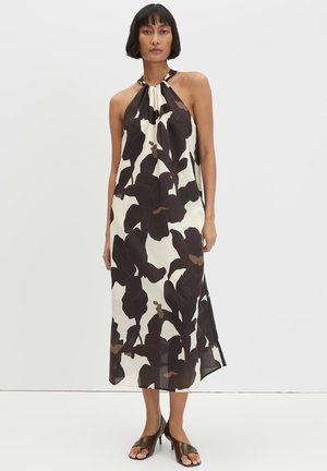 Frau trägt ein ärmelloses Midikleid mit großem schwarzem Blumenmuster auf weißem Hintergrund, kombiniert mit braunen offenen Sandalen mit Absatz.