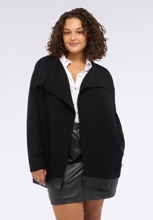 Zwarte oversized cardigan met een geribbelde textuur, open voorkant en zijzakken, gedragen over een witte shirt en een zwarte leren rok.