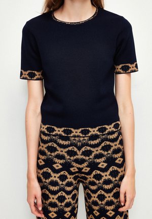 Femme portant un haut noir à manches courtes et un pantalon assorti avec des motifs géométriques et floraux beiges et noirs sur les bords et la partie inférieure.