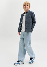 Tengerészkék-fehér kockás, gombos ing, alatta fehér póló, világoskék bő farmernadrág és kék részletekkel díszített sportcipők. Mosolygós póz.