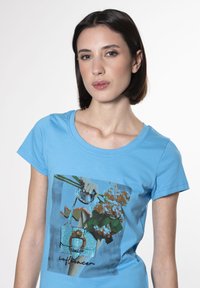 Camiseta de manga corta azul claro con un estampado gráfico de una mano sosteniendo un bolso y flores. Material de algodón suave, escote redondo.