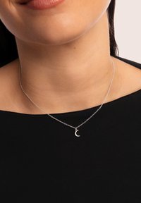 Collier en chaîne argentée avec un petit pendentif en forme de croissant de lune, orné de détails scintillants, porté sur un haut sombre.