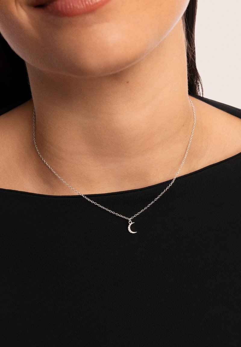 Collier en chaîne argentée avec un petit pendentif en forme de croissant de lune, orné de détails scintillants, porté sur un haut sombre.