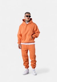 Hoodie orange avec une poche avant et un cordon de serrage, associé à un pantalon de jogging assorti. Les deux articles ont des poignets côtelés et sont fabriqués en tissu doux.