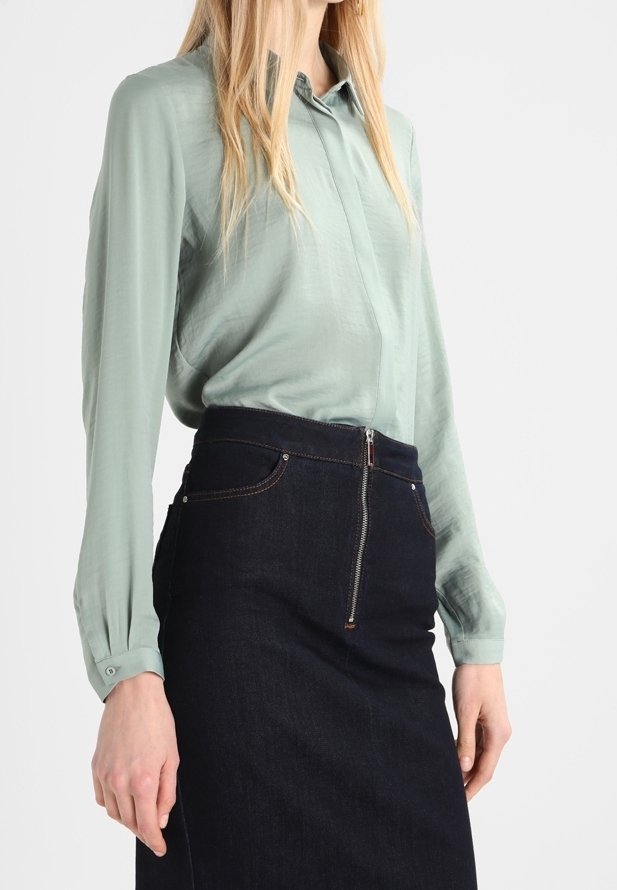 Femme portant un chemisier vert clair à manches longues rentré dans une jupe en denim foncé taille haute avec une fermeture éclair à l'avant.