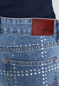 Denim teksad, millel on pruun nahklabel, kaunistatud erineva suurusega läbipaistvate nupukestega, rõhutades tekstuurset, kulunud sinist kangast.