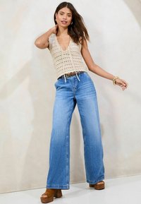 Beige gehäkelter Oberteil mit gewelltem Ausschnitt, kombiniert mit hoch-taillierten, weiten blauen Denim-Jeans und braunen Plateauschuhen.