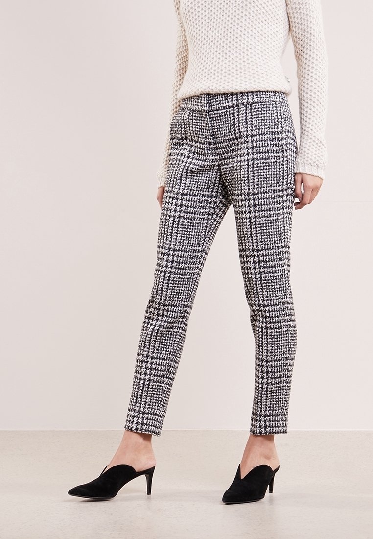 Femme portant un pantalon slim à carreaux noir et blanc, un pull en maille crème et des mules à talons pointus noirs, debout sur un sol clair.