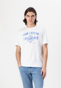 Balta medvilnės marškinėliai su mėlynu grafikos tekstu "TOM TAILOR URBAN FASHION GEAR 1962" ir apskrito logotipo. Trumpomis rankovėmis ir apvaliu iškirpimu.