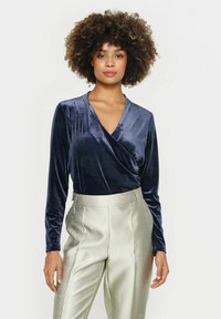 Navy velvet long-sleeve wrap top paired with shiny, light-colored wide-leg trousers. Soft texture with a V-neck design.