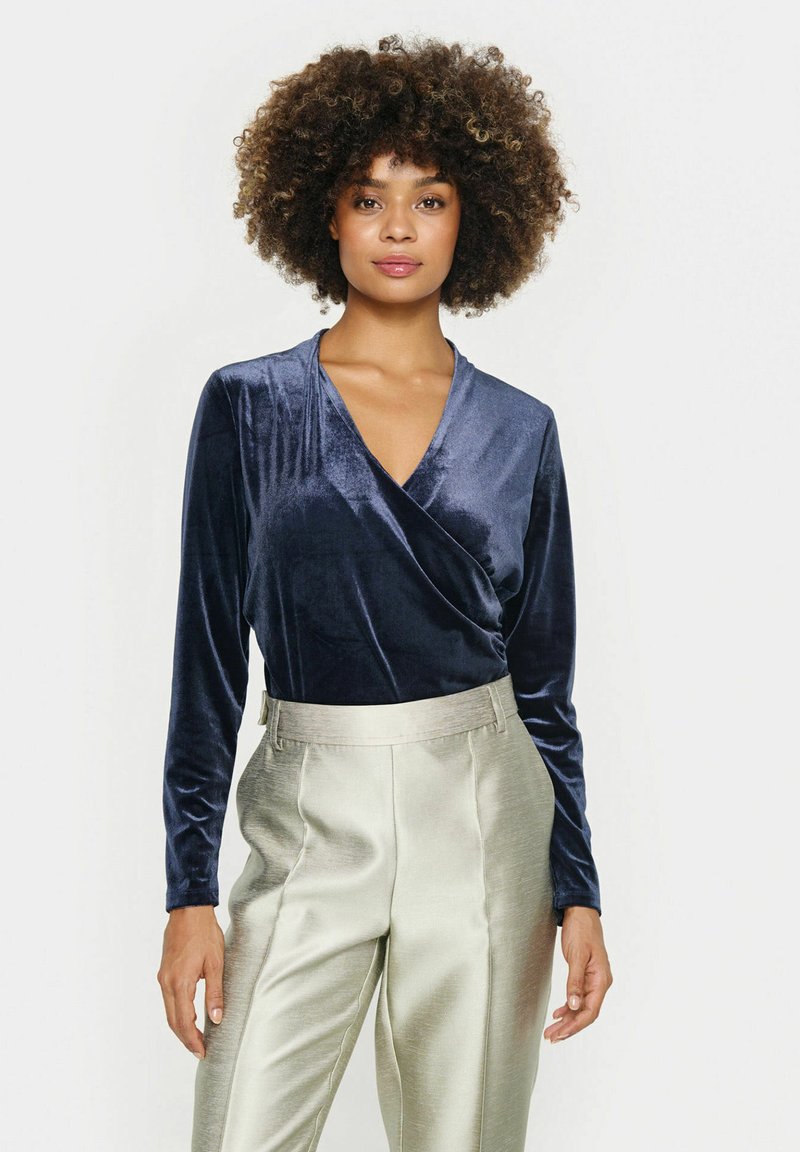Navy velvet long-sleeve wrap top paired with shiny, light-colored wide-leg trousers. Soft texture with a V-neck design.