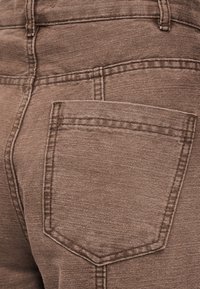 Κοντινή λήψη σε καφέ denim ύφασμα που δείχνει την πίσω τσέπη και τις λεπτομέρειες ραφής ενός ζευγαριού παντελονιών.