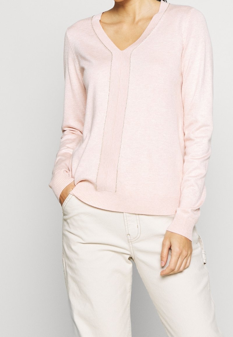 Femme portant un pull léger rose pâle à manches longues avec un col en V et un détail de couture verticale, ainsi qu'un pantalon crème, la main dans la poche.