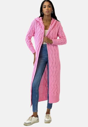 Lange roze gebreide cardigan met kabelgebreide structuur, capuchon en open voorkant. Gecombineerd met nauwsluitende blauwe spijkerbroek en witte sneakers.