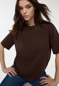 Brązowa koszulka z krótkim rękawem wykonana z materiału bawełnianego, charakteryzująca się luźnym krojem i prostym okrągłym dekoltem. Połączona z niebieskimi dżinsami.