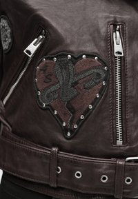 Καφέ δερμάτινο μπουφάν με ένα μπαλώμα σε σχήμα καρδιάς με σπασμένο σχέδιο, ασημένια τρουκ και φερμουάρ στο μπροστινό μέρος. Υφή με λεπτομέρειες και δομημένο σχήμα.