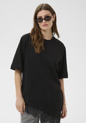 Pikkade pruunide juustega naine, kes kannab musta asümmeetrilist T-särki, halli pitsdetailidega pükse, ovaalseid musti päikeseprille ja kuldseid ehteid.