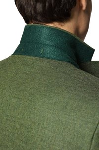 Zielony wełniany płaszcz z wysokim kołnierzem o filcowej fakturze i kontrastującymi ciemnozielonymi akcentami wokół szyi. Widoczna faktura materiału.