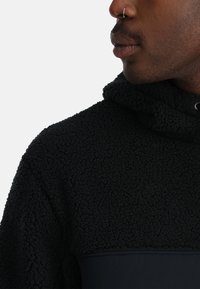 Felpa con cappuccio nera in pile con superficie texturizzata, caratterizzata da un pannello di tessuto solido a contrasto. Il cordoncino regolabile del cappuccio aggiunge funzionalità.