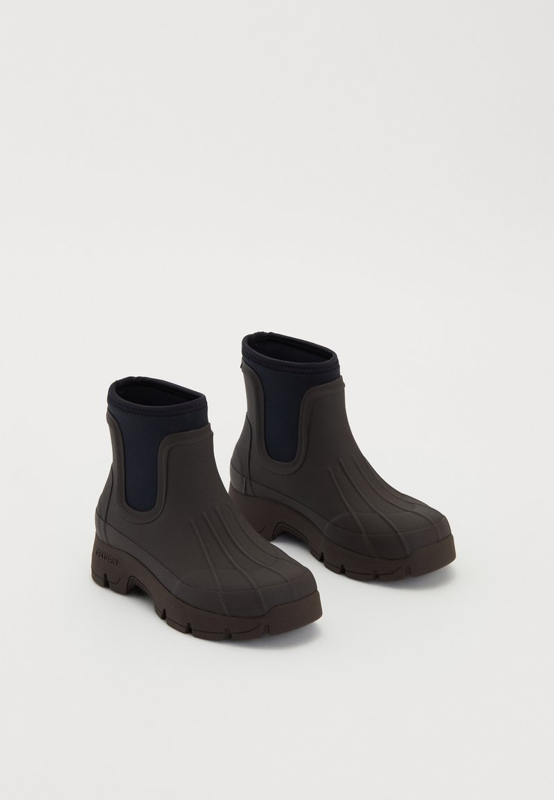 Par de botas de lluvia negras hasta el tobillo con suelas gruesas y paneles laterales elásticos sobre un fondo blanco.