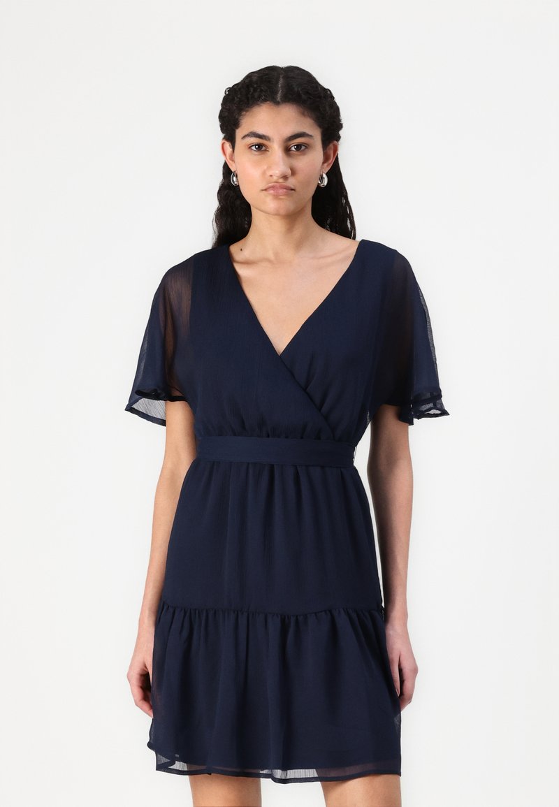 Ναυτικό μπλε φόρεμα τύπου wrap από ελαφρύ ύφασμα, με κοντά φτε�ρωτά μανίκια, εφαρμοστή μέση και φουσκωτό στρίφωμα.