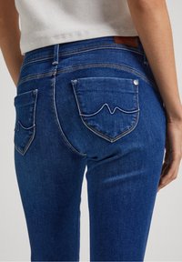 Blå denimjeans med en figurnära design, två bakfickor med ett vågigt sömmönster och en läderetikett på midjebandet.