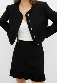 Černé saka s tvarovaným výstřihem, uzavřením na zlaté knoflíky a přední kapsou, spárované s černou skládací sukní.