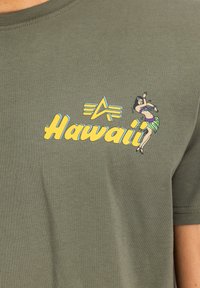 Oliven grøn T-shirt med gul "Hawaii" tekst, en grafik af en hula danser og blå accentlinjer. Fremstillet af blødt bomuldsstof.