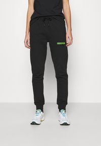 Pantalones de chándal negros con un corte ajustado, que cuentan con una cinturilla elástica con cordón y un parche de logo verde en el muslo. Tejido suave, con un detalle mínimo.
