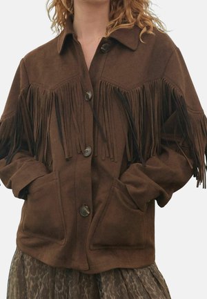 Femme portant une veste en daim marron boutonnée avec des détails à franges sur la poitrine et le haut des bras, les mains dans les poches avant.