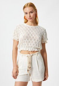 Top corto de ganchillo blanco con mangas cortas abullonadas, un dobladillo con volantes, combinado con pantalones cortos de talle alto en beige claro que cuentan con un cordón trenzado.
