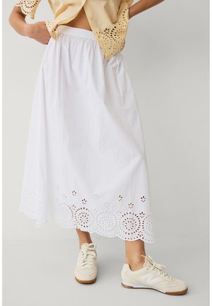 Kobieta ubrana w białą spódnicę midi z kwiatowym wykończeniem ażurowym oraz beżowe sneakersy, w komplecie z beżową bluzką z krótkim rękawem i ażurowym wzorem.