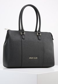 Czarna torebka skórzana o fakturze, z dwoma uchwytami i złotym logo Armani Jeans na przodzie, umieszczona na białej powierzchni na tle jednolitego tła.